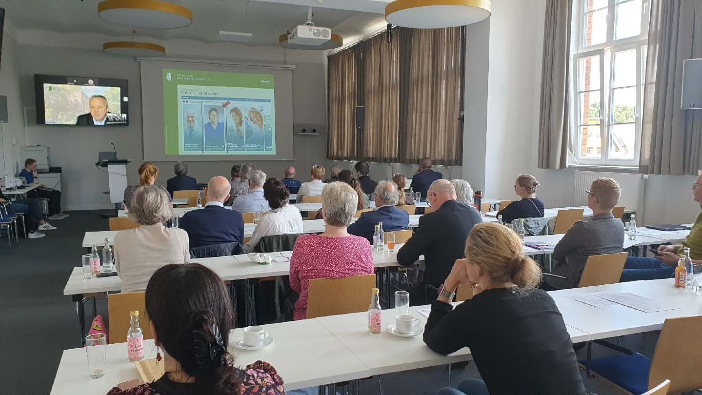 View into the conference room: participants sit at rows of tables; at the front a presentation and a speaker joining by video can be seen.